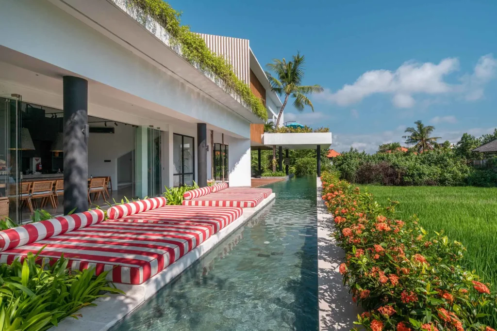 A luxurious tropical villa named Mandala Oasis Villa, featuring an infinity pool surrounded by palm trees, lush gardens, and modern open-air architecture under a bright blue sky.