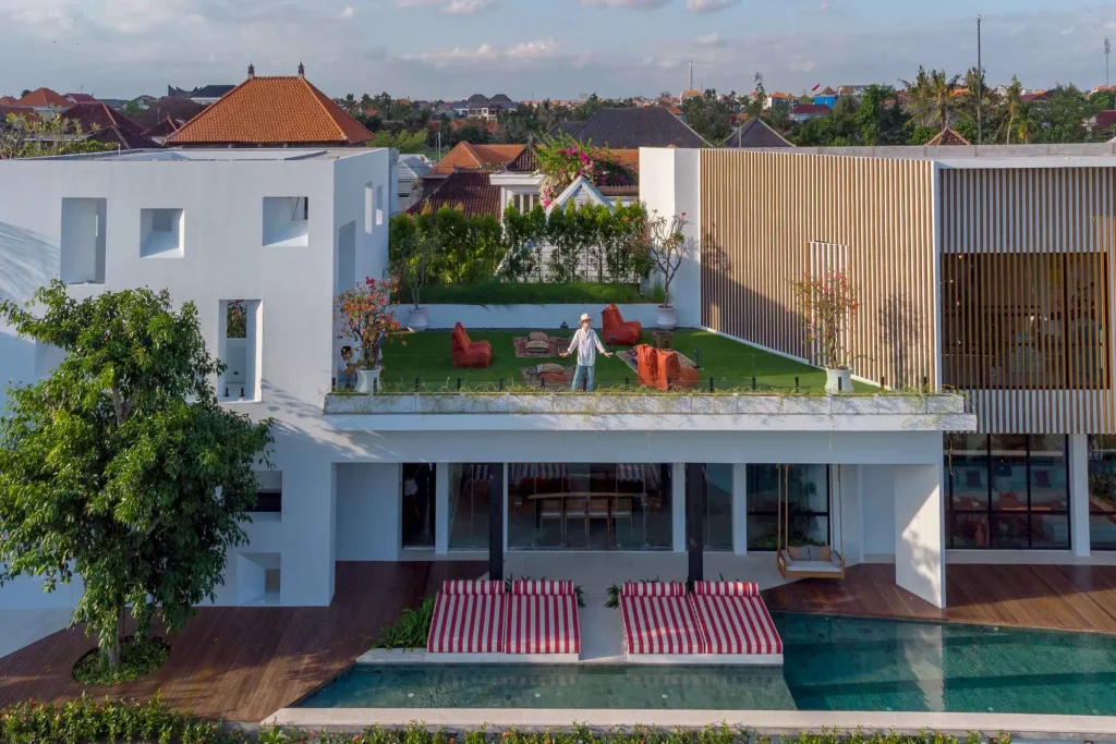 A luxurious tropical villa named Mandala Oasis Villa, featuring an infinity pool surrounded by palm trees, lush gardens, and modern open-air architecture under a bright blue sky.